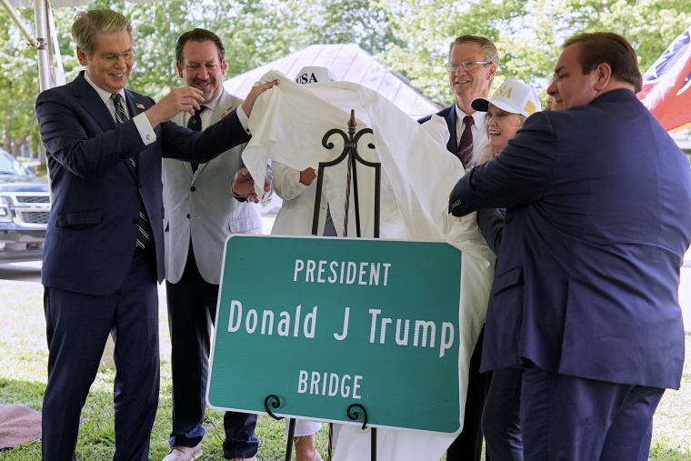 Scott Bessent and other officials pull a white sheet off a green roadsign reading PRESIDENT DONALD J TRUMP BRIDGE